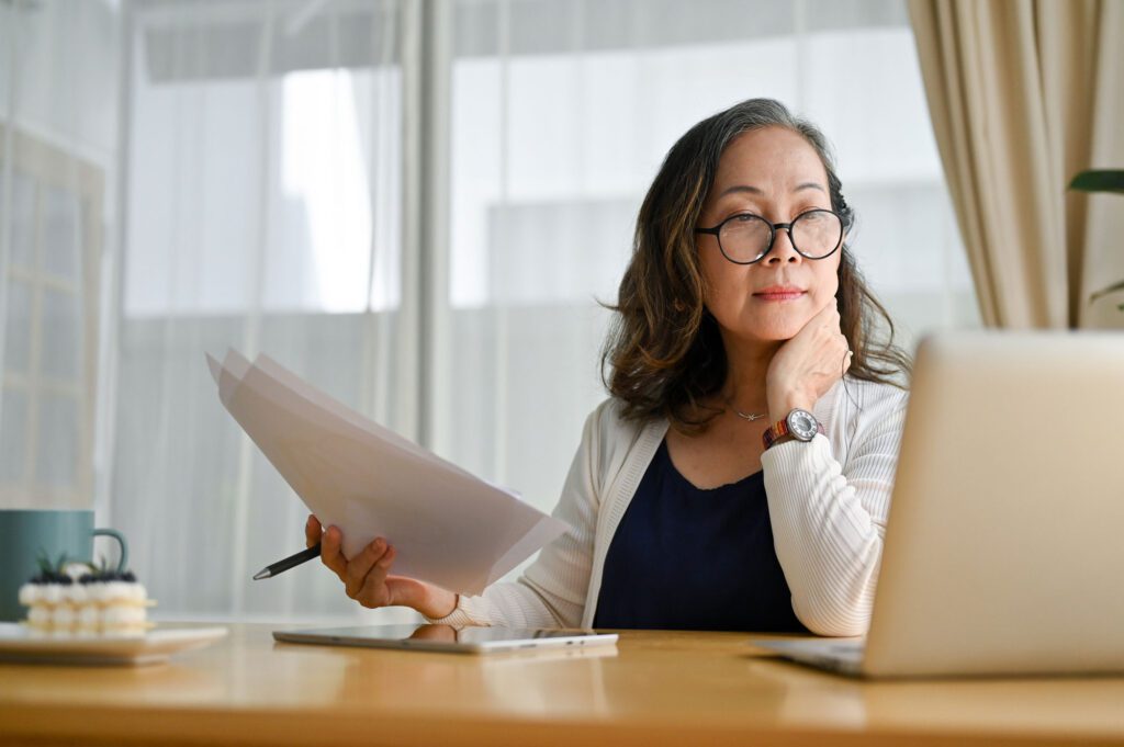 Concentrated asian middle aged female teacher in glasses sitting at desk using portable computer and examining paperwork