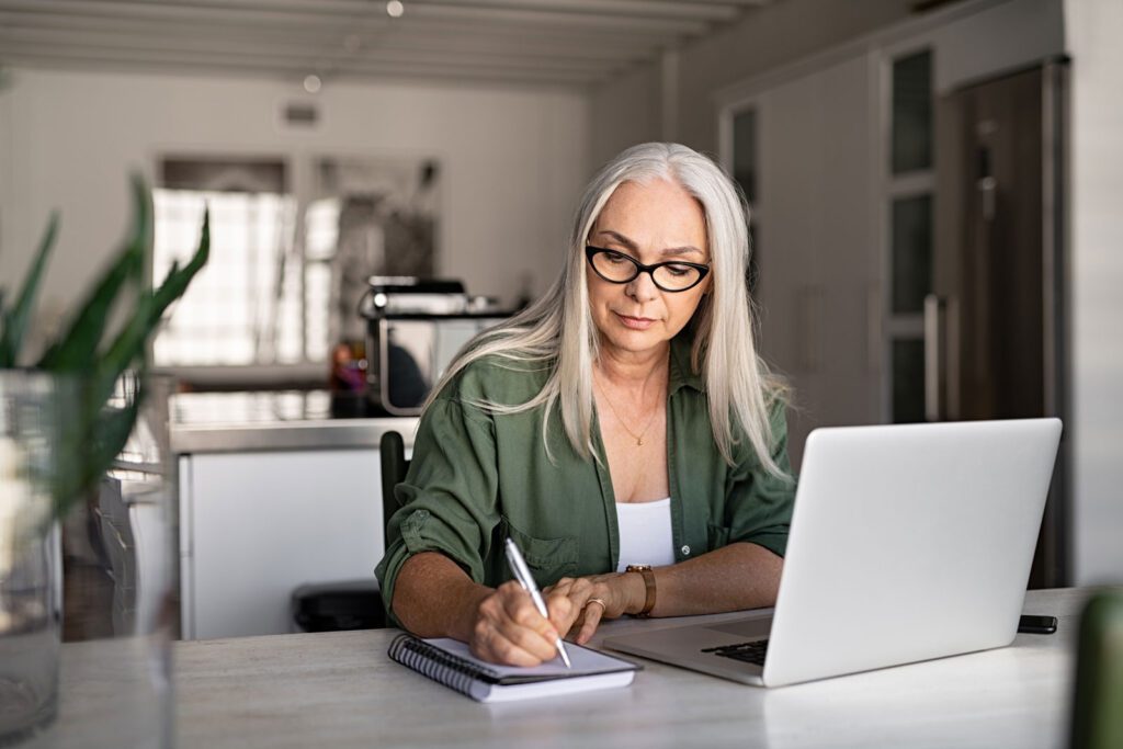 Senior stylish woman taking notes in notebook while using laptop at home